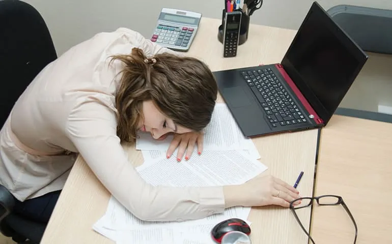 Woman resting her head on a desk next to a laptop, looking exhausted