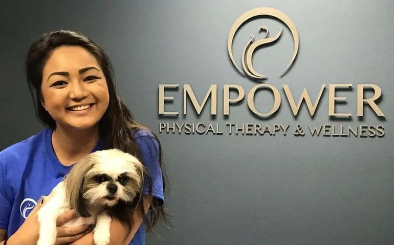 Woman holding a small dog in front of an Empower Physical Therapy & Wellness backdrop