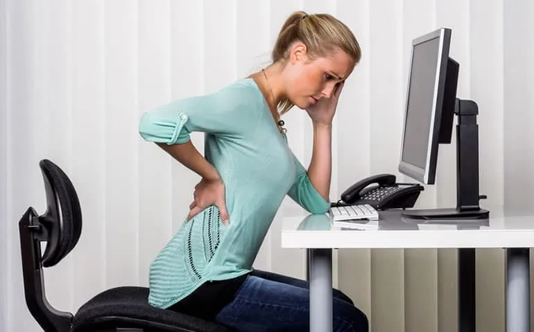 Woman standing at a desk holding her lower back in pain